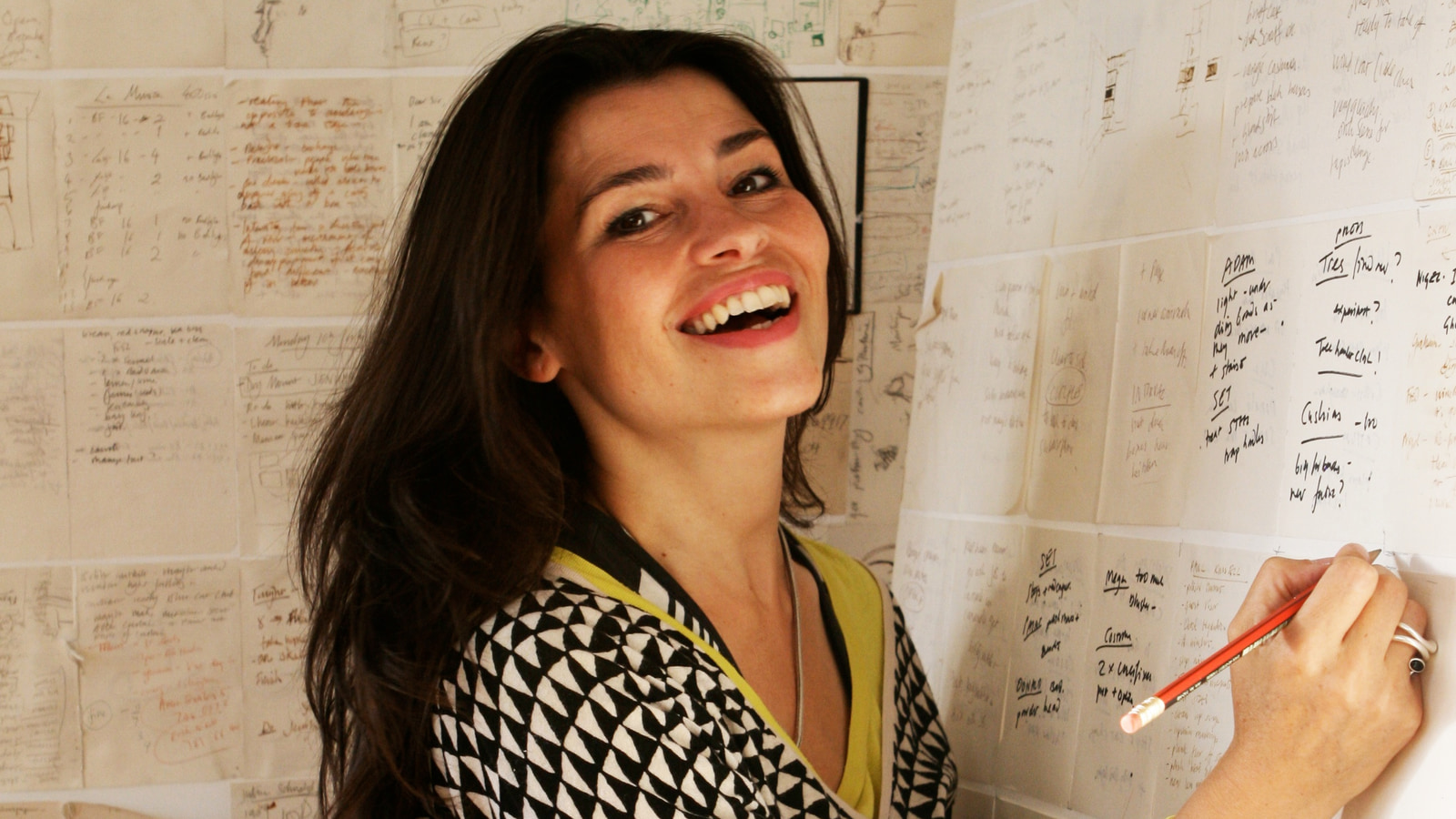 Es Devlin smiling in her studio, holding a pencil beside a wall of handwritten notes.